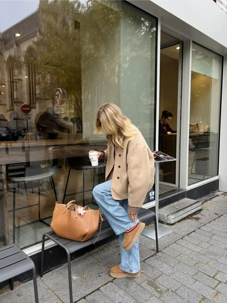 Woman in a camel jacket and wide-leg jeans stepping onto a bench outside a café