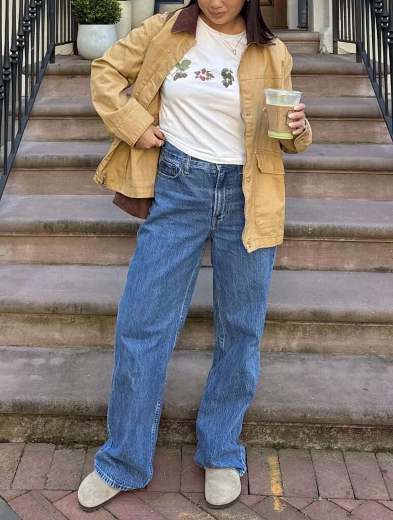 Woman in a tan chore jacket and white graphic tee standing on outdoor steps holding a drink