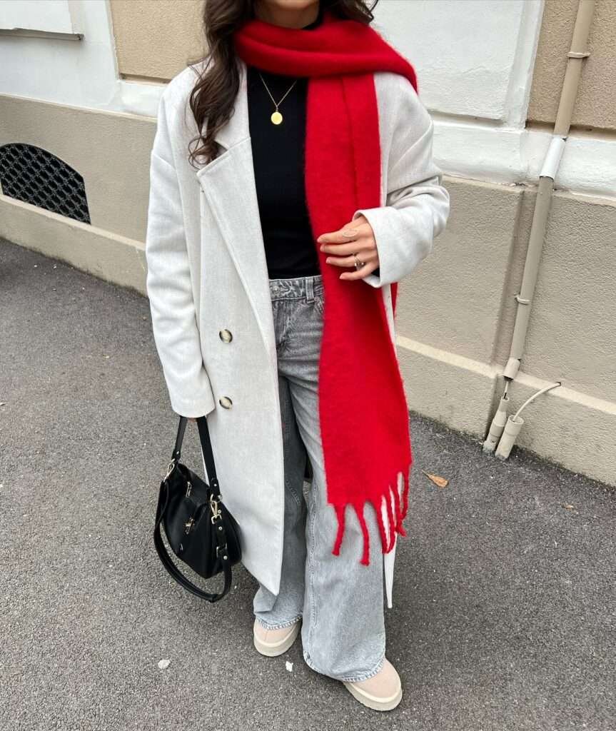 Woman wearing an oatmeal wool coat with a bright red fringed scarf over grey wide-leg jeans