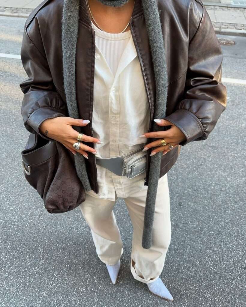 Close-up of a leather bomber jacket worn over cream linen layers with a waist bag and layered rings