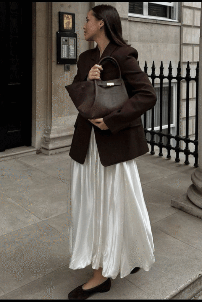 Woman in an oversized brown blazer and voluminous ivory satin skirt walking outside a townhouse