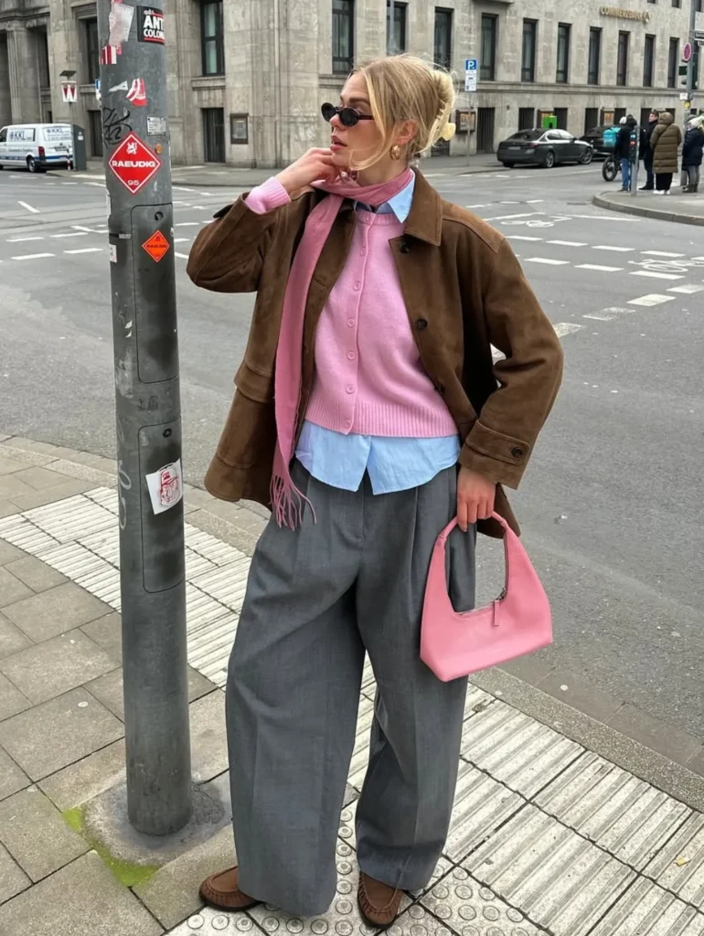 Woman in a brown suede jacket and pink cardigan standing by a street lamp holding a pink bag
