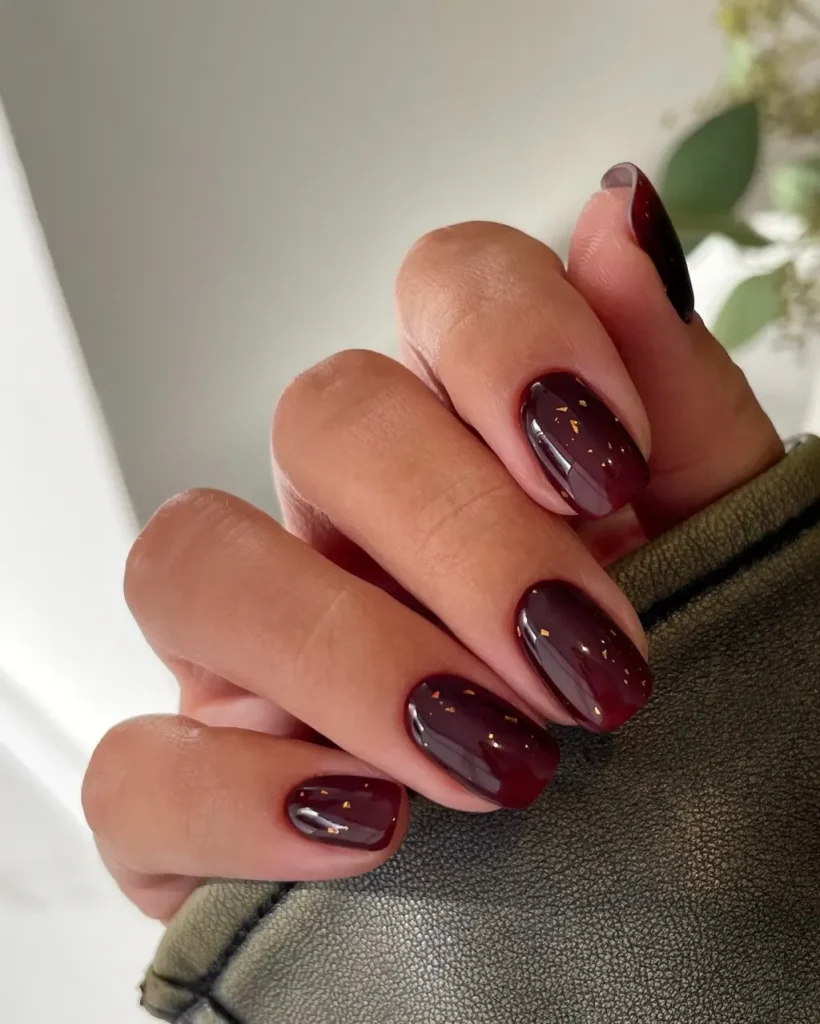 Short-to-medium oval nails in a glossy oxblood polish with scattered gold foil flakes.