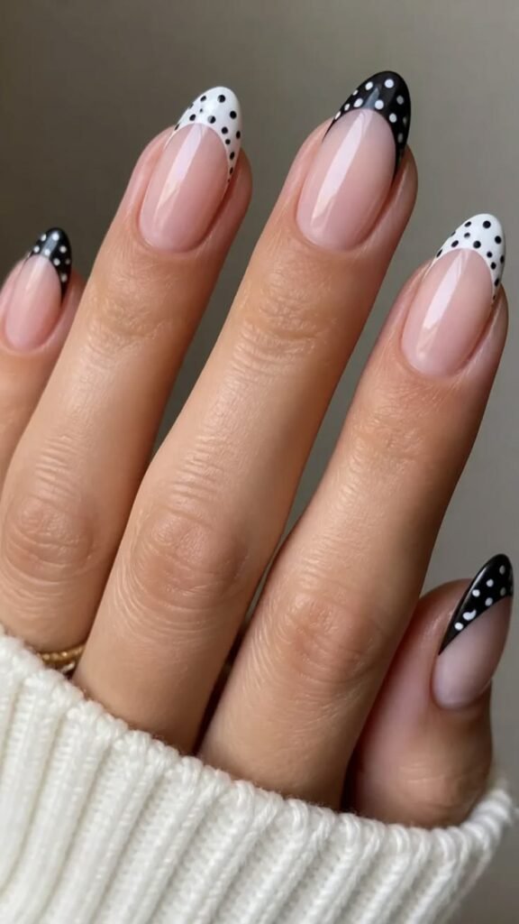 Almond-shaped nails with a sheer nude base and alternating black-and-white polka dot French tips, worn with a cream sweater sleeve.