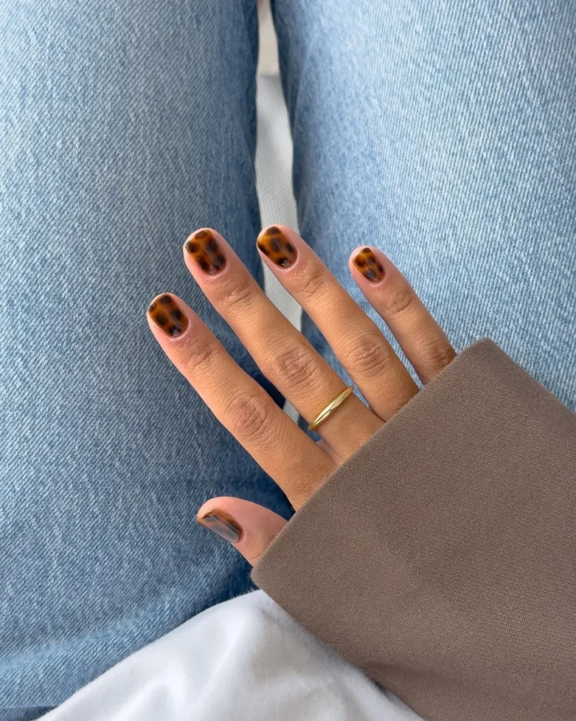 Short square nails painted in a brown and black tortoiseshell pattern, worn with denim.