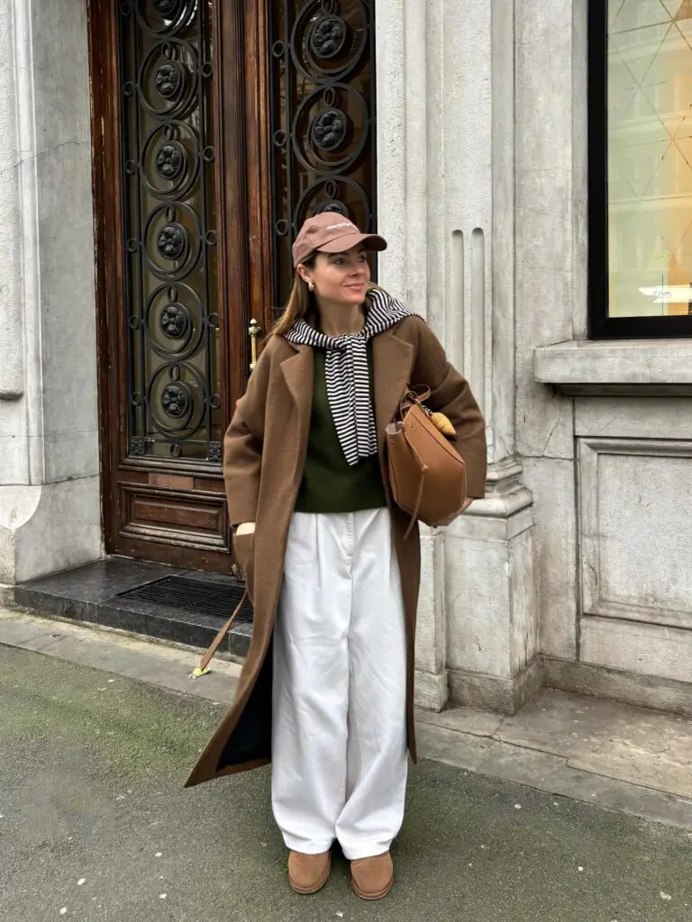 This ensemble effortlessly pairs relaxed street style with rich, elevated tailoring. A full-length, chocolate-brown wool trench coat acts as the anchor, giving the outfit instant structure and presence. Beneath it, a forest green knit offers an understated color-blocking moment paired against crisp white, wide-leg trousers. The striped knit draped loosely over the shoulders adds a playful Parisian flair, while the baseball cap and suede boots ground the look in laid-back elegance. It’s the ultimate blueprint for cool-girl off-duty layering.