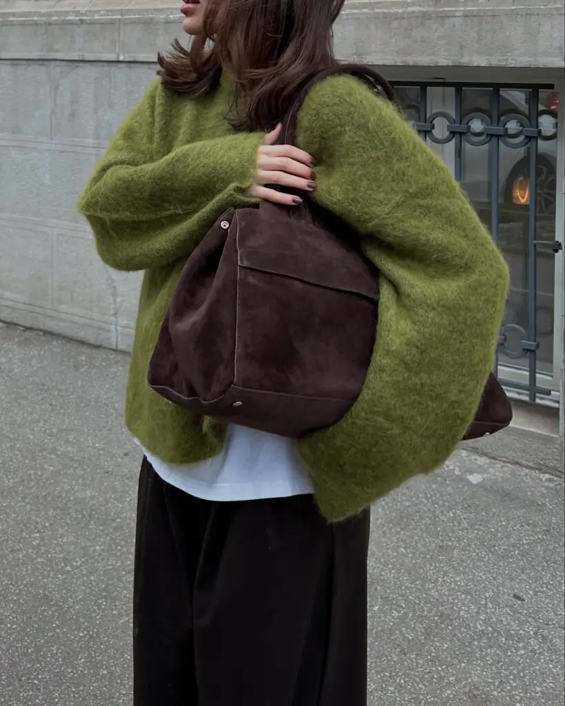Tactility takes center stage here with an oversized, fuzzy mohair knit in a striking olive shade. Layered over a simple white tee, the contrast of smooth cotton against rich wool adds a subtle sense of depth. Dark, wide-leg trousers maintain the easygoing silhouette while keeping the focus firmly on the statement knit. The look is beautifully finished with a slouched espresso-suede tote bag that mirrors the autumn-inspired tones. It’s minimalist dressing done with maximum texture.