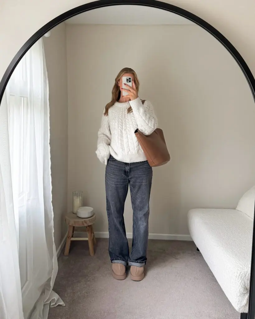 This outfit proves that a great cable-knit sweater is the cornerstone of any cold-weather wardrobe. The rich, braided texture of the cream knit adds timeless depth to a clean, understated palette. Paired with relaxed charcoal grey jeans, the look hits the ideal balance between casual and thoughtfully put-together. A structured tan leather bucket bag introduces a warm, tonal contrast that ties in with plush suede boots. It’s effortless, everyday chic executed to perfection.
