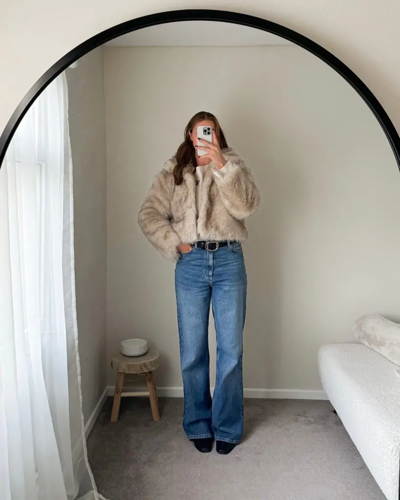 Cropped plush faux fur gets a casual reinterpretation when paired with classic, full-length denim. The soft, sandy-beige jacket brings immediate visual softness and warmth to the upper half. Tucking a lightweight knit into high-rise jeans defines the waist, highlighted by a clean black leather belt with gold hardware. Pointed dark boots peeking out under wide hems elevate the entire profile from simple to sharp. It’s a fresh, modern take on daytime dressing.