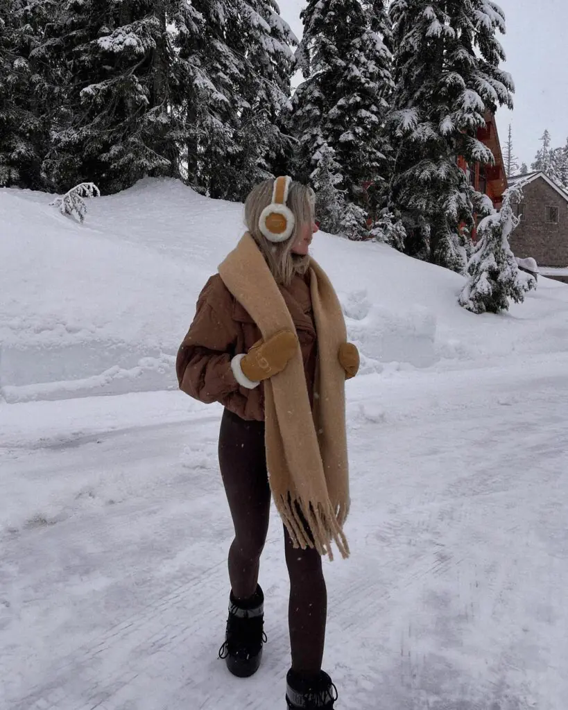 Designed for cold-weather adventures, this ensemble combines high-function gear with thoughtful color harmony. A quilted brown bomber jacket pairs seamlessly with matching deep-brown leggings for a streamlined base. The oversized, thick-fringe camel scarf and matching UGG earmuffs infuse the outfit with ultra-cozy appeal. Heavy-duty black snow boots anchor the look, making it completely practical for snowy landscapes. It’s a seamlessly coordinated alpine wardrobe moment.