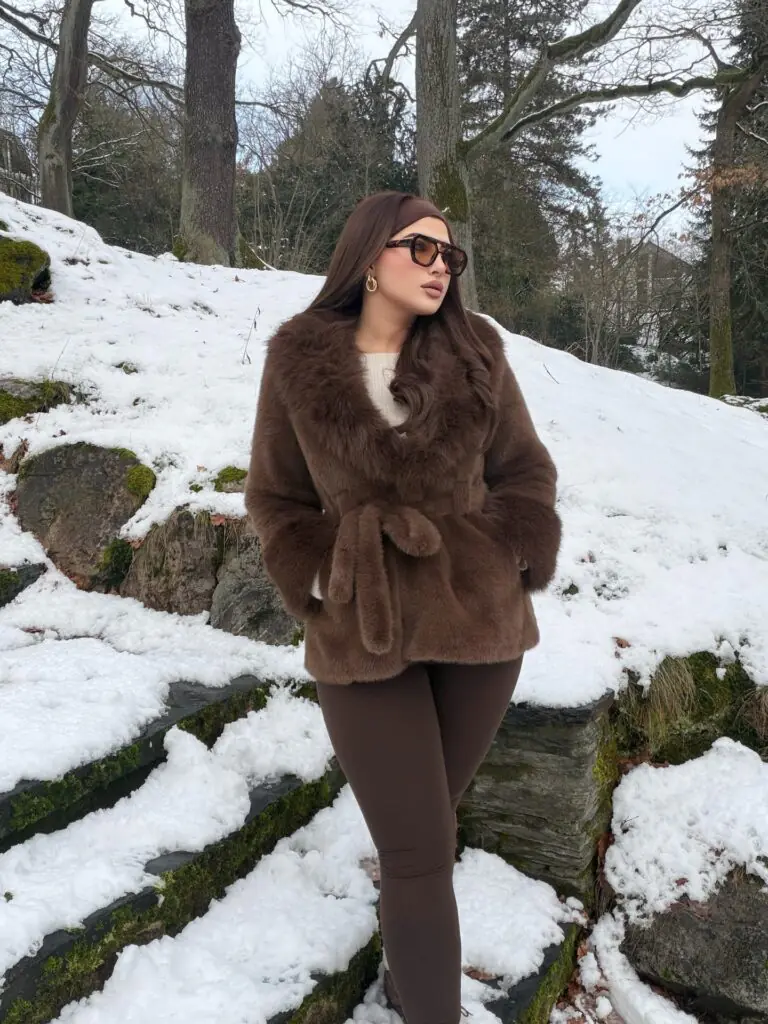 Rich chocolate tones create an undeniably luxurious, monochromatic aesthetic in a snowy setting. The focal point is a lush, belted faux-fur jacket featuring a wide shawl collar that frames the face beautifully. Cinching the waist adds definition and elegance to an otherwise voluminous winter piece. Matching dark brown leggings keep the bottom sleek, letting the statement coat steal the spotlight. Golden hoop earrings and tinted tortoiseshell sunglasses add the final touches of vintage glamour.