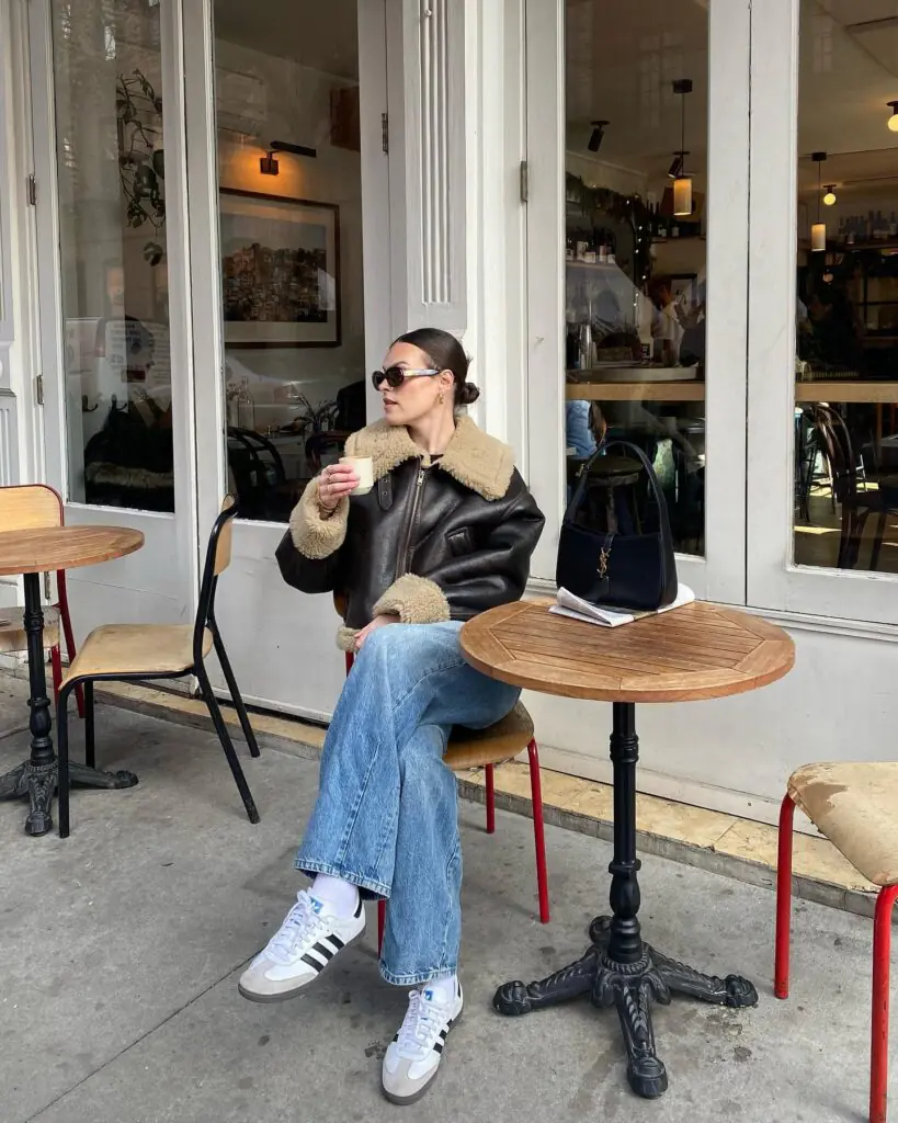 The image captures an off-duty street-style archetype composed of a dark leather shearling aviator jacket featuring plush contrast fleece cuffs and an oversized collar, styled alongside relaxed light-wash straight-leg denim, white terrace sneakers with crew socks, black oval sunglasses, and a structured black leather shoulder bag.