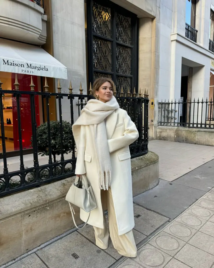 This look showcases a fully monochromatic cream ensemble featuring a double-breasted maxi wool coat, an oversized brushed knit scarf wrapped around the neck, fluid wide-leg cream tailored trousers, and a soft ecru leather tote bag, set against a Parisian street backdrop.