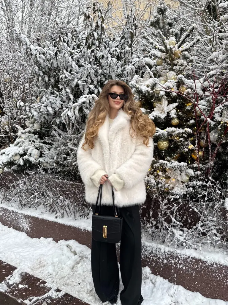 Set against a snow-covered alpine landscape, this image displays a mid-length, plush white faux fur coat paired with fluid black wide-leg trousers, a structured black crocodile-embossed leather tote bag, dark cat-eye sunglasses, and voluminous styled waves.