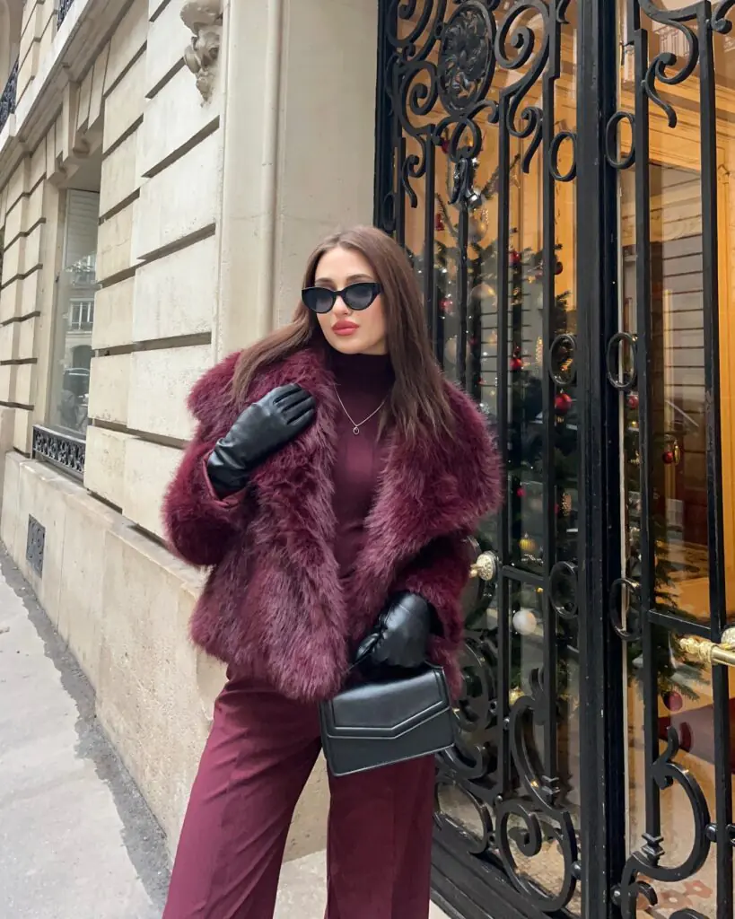 This monochromatic look features a shaggy deep burgundy/oxblood faux fur jacket worn over a color-matched turtleneck sweater and tailored burgundy trousers, complemented by sleek black fitted leather gloves, a structured black handbag, and black cat-eye sunglasses.
