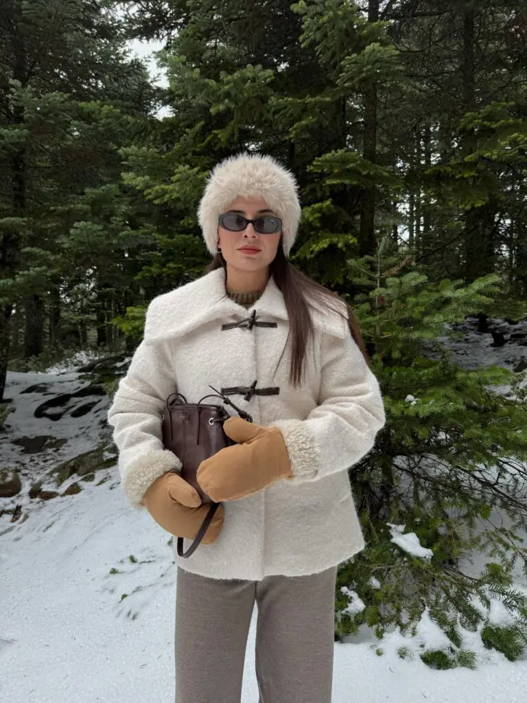 This image presents a winter alpine aesthetic set in a snowy pine forest, featuring an ivory wool-and-shearling jacket with dark brown leather toggle closures, an oversized collar, a plush furry pillbox hat, tan suede-and-fleece mittens, and a dark brown leather bucket bag.