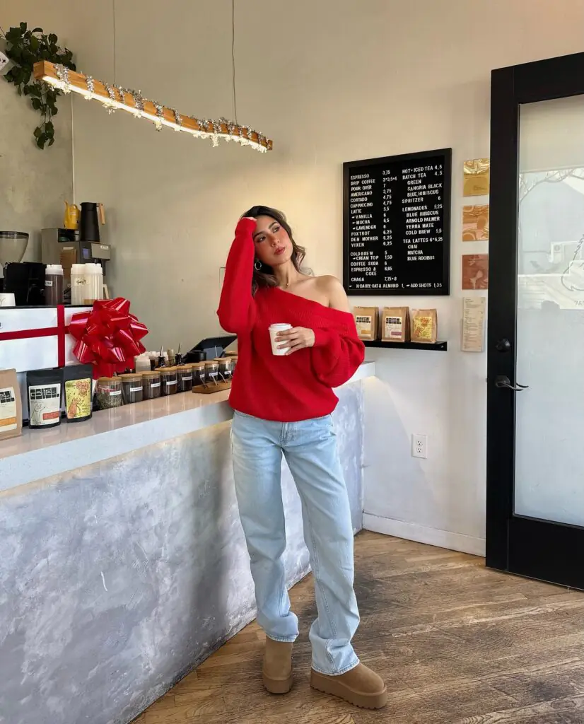 Captured inside a coffee shop, this casual look consists of an asymmetric, off-the-shoulder bright cherry red sweater paired with light-wash relaxed denim, platform tan suede Ugg-style boots, and minimal silver jewelry.