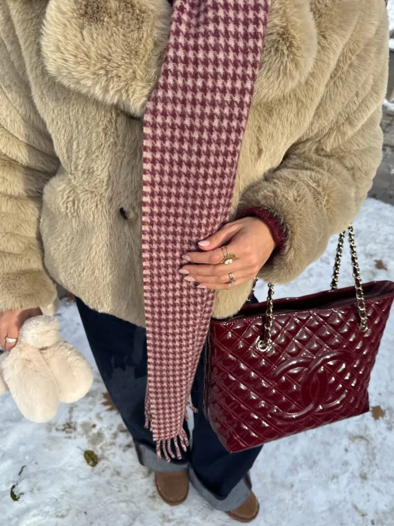 This close-up outdoor detail shot features a plush beige/tan faux fur jacket layered with a woven burgundy-and-cream houndstooth wool scarf, a glossy burgundy quilted patent leather Chanel bag with chain straps, and tan suede footwear.